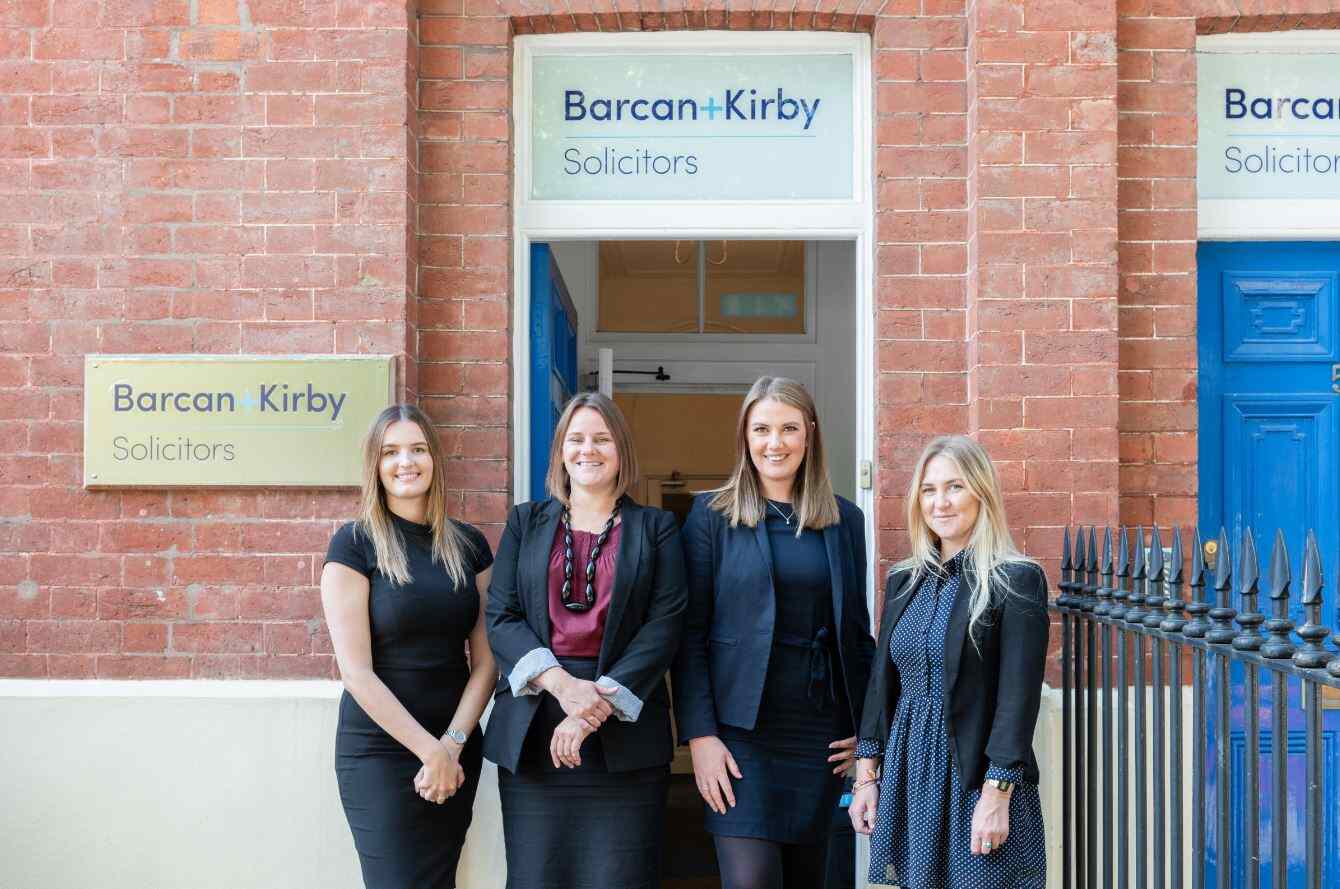 Four smartly dressed young females in front of a red brick office with the Barcan and Kirby logo above the door