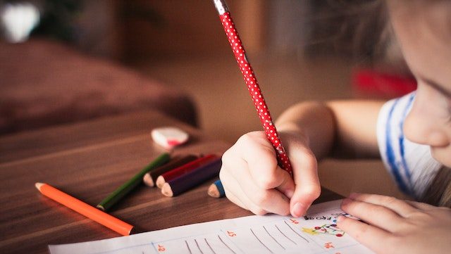 A young child writing on a sheet of paper using a red pencil with white polka dots. Several coloured pencils are lying on the table nearby.