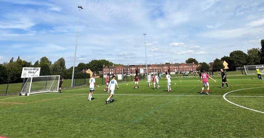 A group of young people in football kits playing on a green football pitch against blue sky