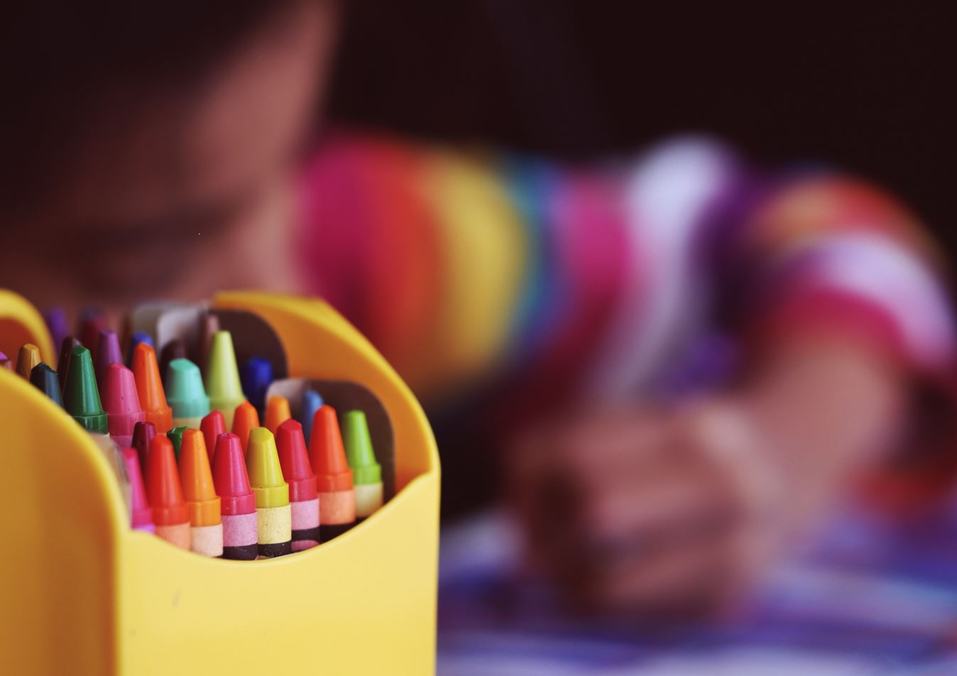 Blurred image of child colouring in next to yellow pot of assorted crayons