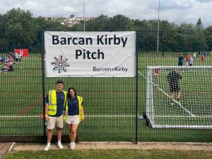 A dark haired male and female dressed in navy blue t-shirts, beige shorts and yellow hi-vis jackets standing in front of a football pitch. There is a large white sign above them that reads 'Barcan Kirby Pitch'.