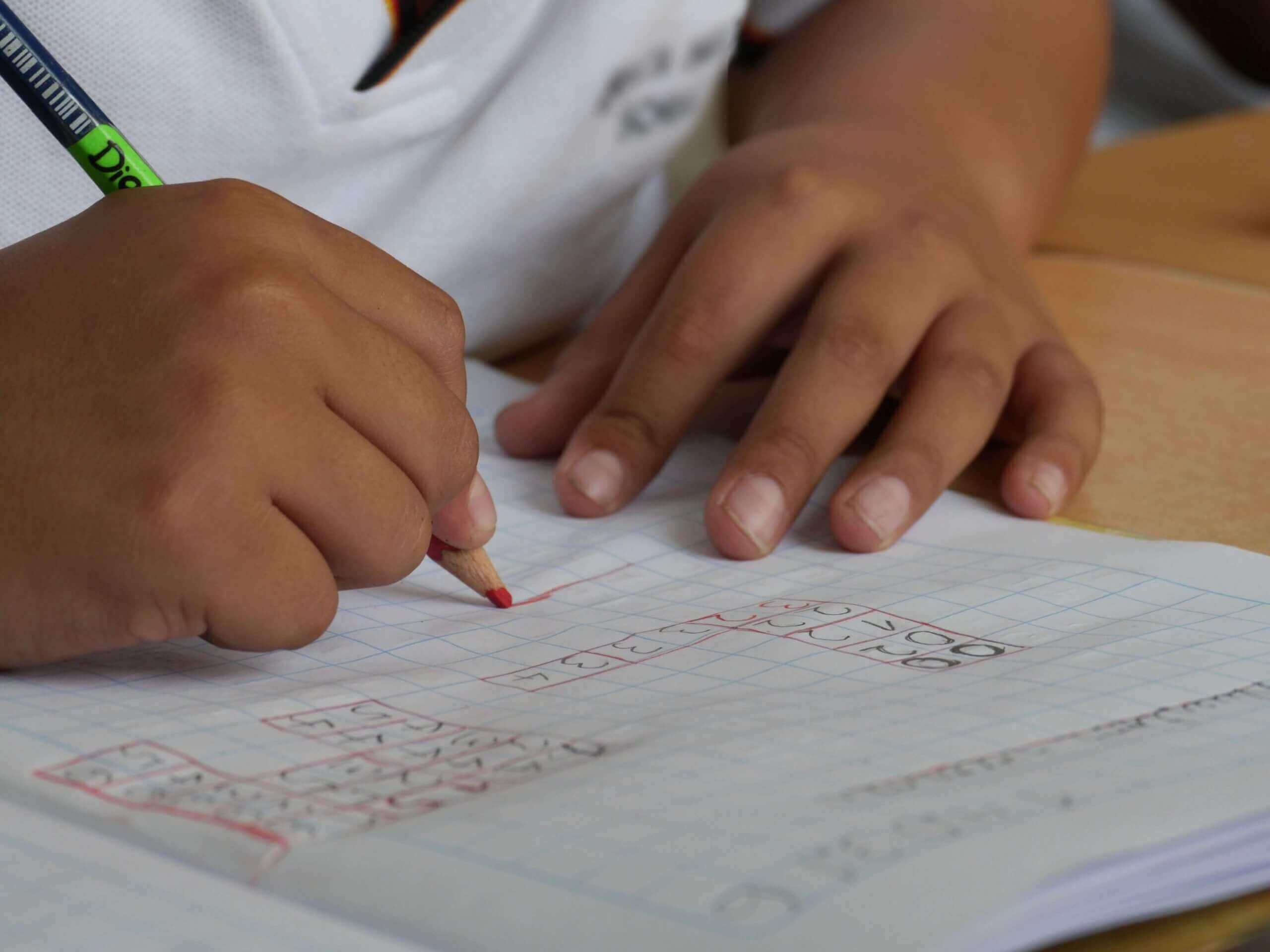 Close-up of a child's holding a red pencil writing or drawing in a notebook with a grid pattern. The background shows a desk and the child's clothing, which is a white shirt.