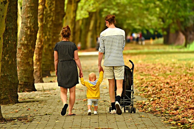 A family of four holding hands and walking together outdoors in a grassy field. The adults are on the outside, holding one child's hand and the other is in a pushchair