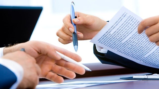 Close-up of four hands engaged in a business meeting. One person is holding and pointing at a document with a pen, while another hand is extended as if gesturing or receiving the pen.