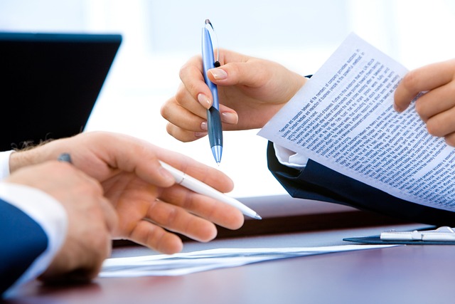 Close-up of four hands engaged in a business meeting. One person is holding and pointing at a document with a pen, while another hand is extended as if gesturing or receiving the pen.