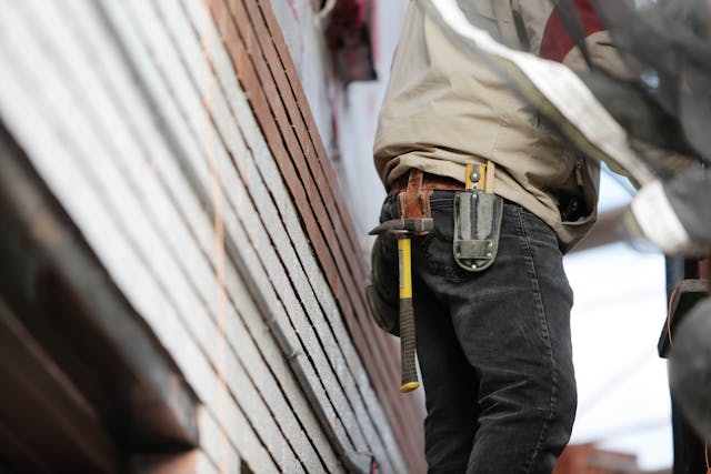 A male construction worker from the waist down, standing on a worksite. He is wearing a tool belt that holds a hammer and other tools, with a background of stacked bricks and building materials.