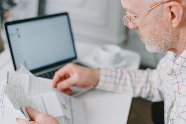 A white ageing man with glasses sitting at a table, holding a stack of documents in one hand while using his other hand to write on a piece of paper.