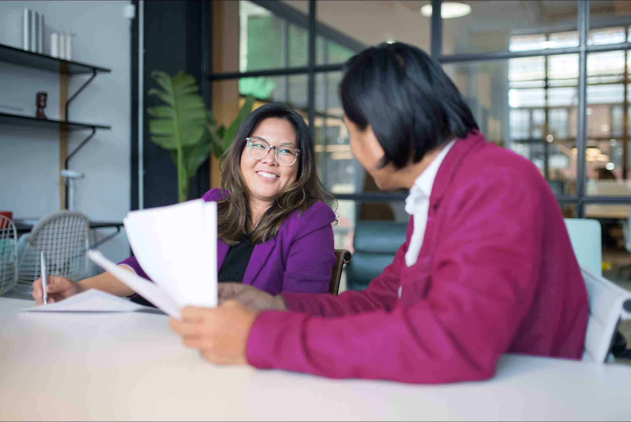 A male and female seated at a table in a modern office setting, engaged in a friendly conversation. The woman on the left is wearing glasses and a purple blazer, while holding a pen and paper. The male on the right also wears a purple blazer and is holding documents.