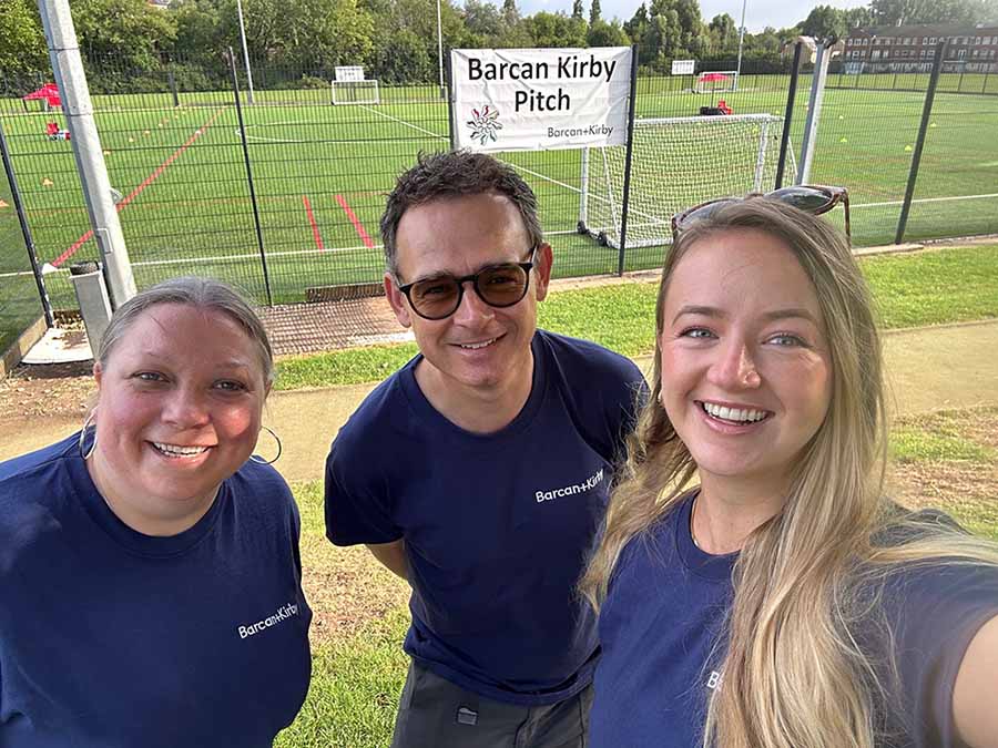 Two blonde haired females either side of a dark haired male who is wearing sunglasses. They are all smiling at the camera, wearing navy blue t-shirts, with a football pitch in the background