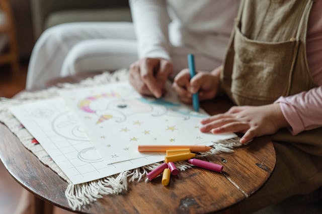 Close-up of an adult and child drawing together at a wooden table, using colourful markers to colour a drawing of a unicorn with stars.