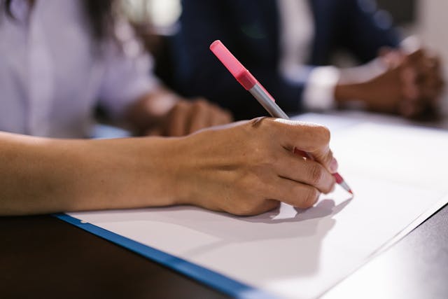 A close-up of a hand holding a pink pen, writing on a piece of paper. The person appears to be seated at a table, with another individual partially visible in the background.