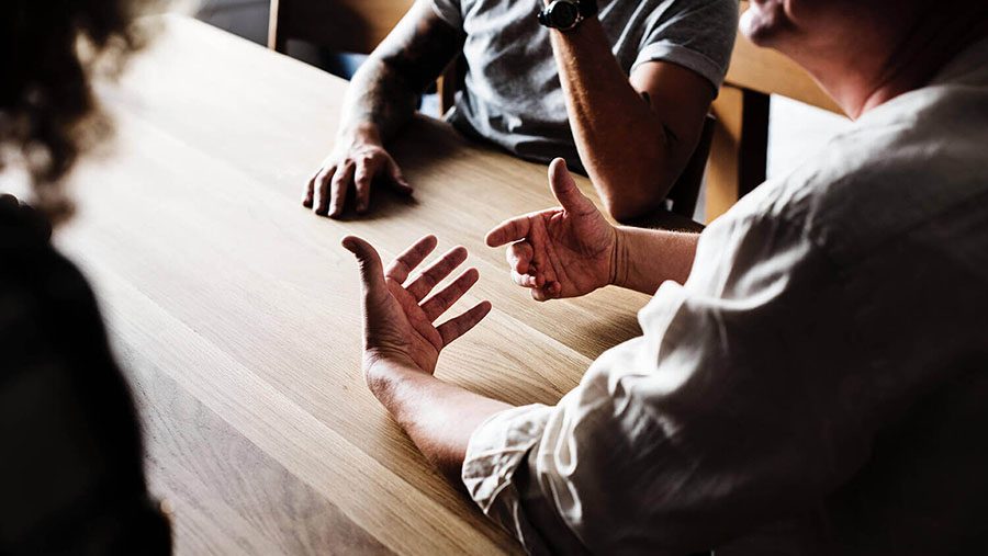 Close-up of three people seated at a wooden table, engaged in a discussion. One person's hands are gesturing expressively while others listen.