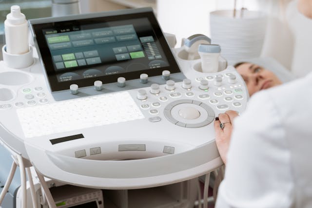 Close-up of an ultrasound machine operated by a healthcare professional. A patient lies in the background, partially visible.