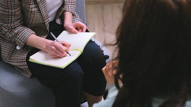 A close-up of two people sitting opposite eachother, with one person noting something down in a notepad