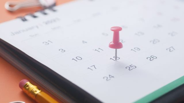 A close-up of a calendar page with a pink push pin, pencil, and binder clip on an orange background