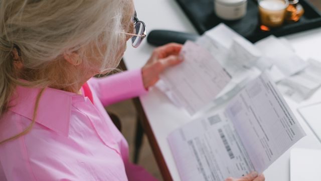 An elderly woman in glasses holds and reads important papers at a table indoors